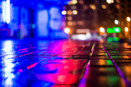 Rainy night in the big city, light from the night club and the windows of the house is reflected in the asphalt. View from the sidewalk level paved with bricks