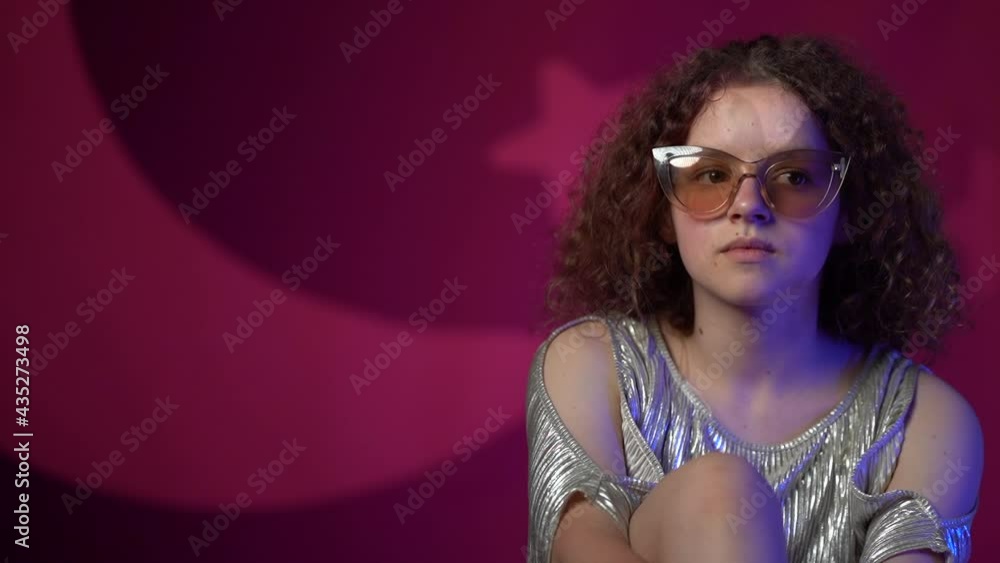 Portrait of a lovely curly girl with stylish glasses in neon lights in a disco club. The teenager is wearing a shiny dress. Studio shooting Portrait of a lovely curly girl with stylish glasses in neon lights in a disco club. The teenager is wearing a shiny dress. Studio shooting