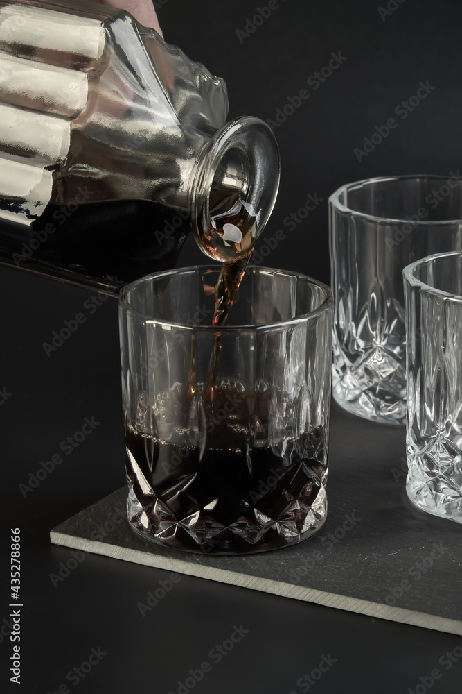 Someone is pouring cognac from a square decanter into a glass next to other empty cognac and whiskey glasses against a dark background. Close-up