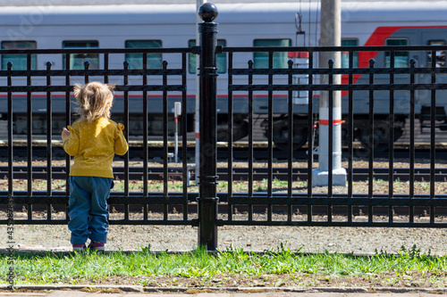A child stands at the fence and looks at a passing train. The child is holding onto the iron fence and looking at the train.