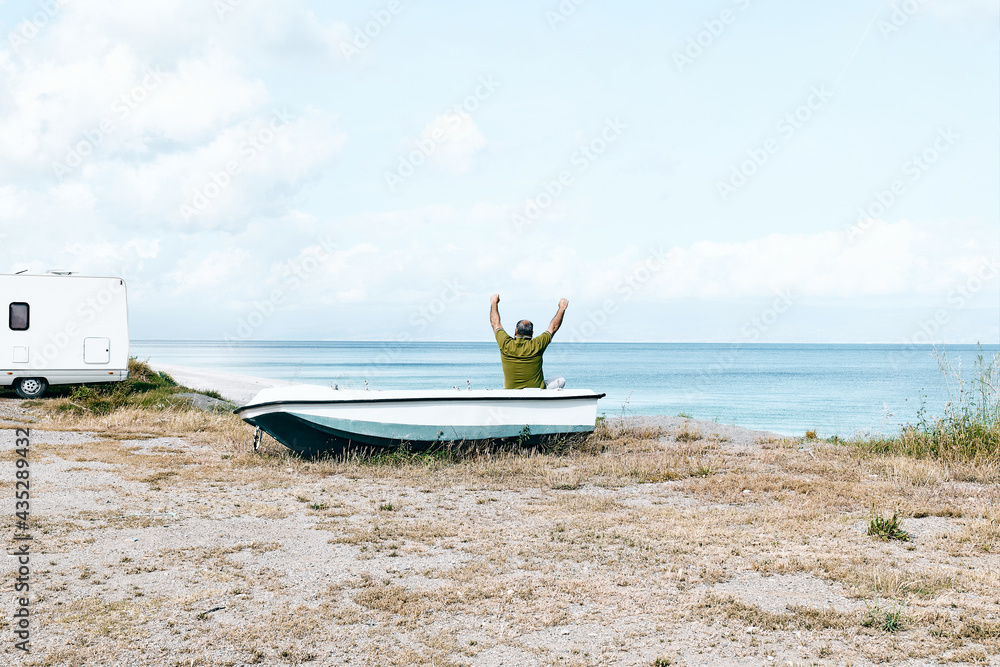 Man sitting in boat on the beach near his white camper van parked by the sea. Tourist season on the mediterranean sea. Sicily. Ionian Sea. Man sitting in boat on the beach near his white camper van parked by the sea. Tourist season on the mediterranean sea. Sicily. Ionian Sea.