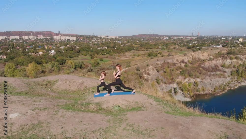 Aerial orbiting shot of slender young mother do yoga exercises with child daughter on high hill by lake in morning, far from bustle of city, set good example and instill in child healthy habits Aerial orbiting shot of slender young mother do yoga exercises with child daughter on high hill by lake in morning, far from bustle of city, set good example and instill in child healthy habits