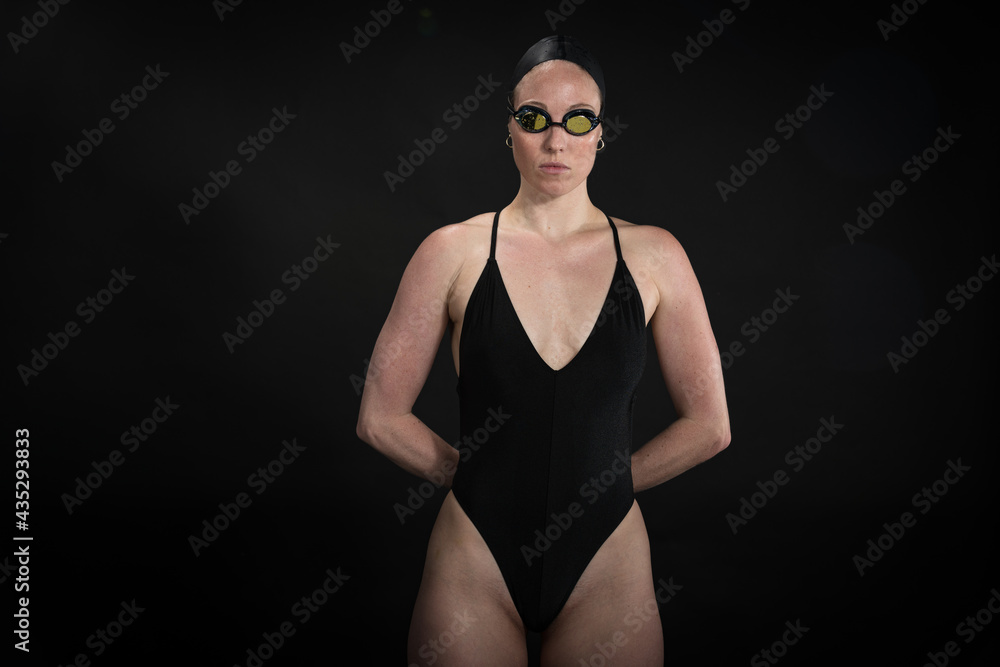 Obraz premium Professional swimmer in suit, cap and goggles poses before a black background.