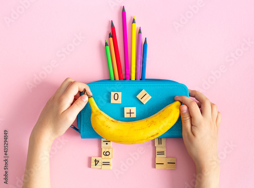 Multicolored school supplies, stationery and a banana in the form of a smiling character in the hands of a child on a pink background. Back to school, early childhood education. School lunch. Flat lay