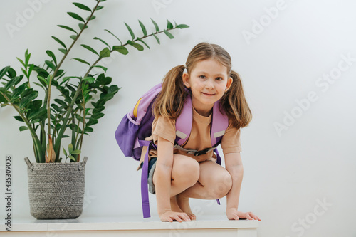 Playful little schoolgirl on a white table with purple backpack. Over white