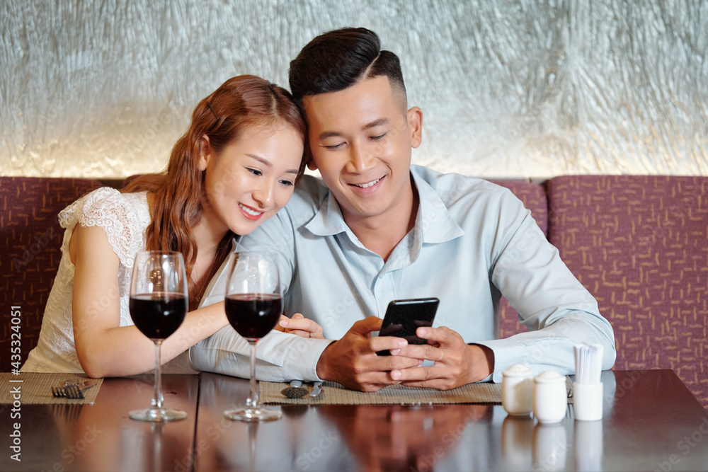 Pretty smiling young woman leaning on shoulder of boyfriend and watching funny video on smartphone when waiting for food in restaurant Pretty smiling young woman leaning on shoulder of boyfriend and watching funny video on smartphone when waiting for food in restaurant