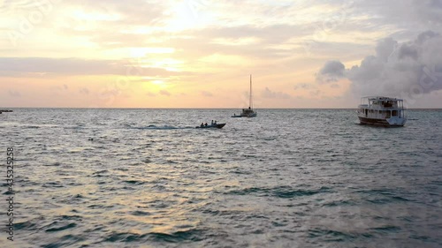 Aerial Moving Forward Towards A Setting Sun As Small Motor Boats Pass By Across Tropical Waters Under Partial Cloudy Sky - Kandooma Fushi, Maldives photo