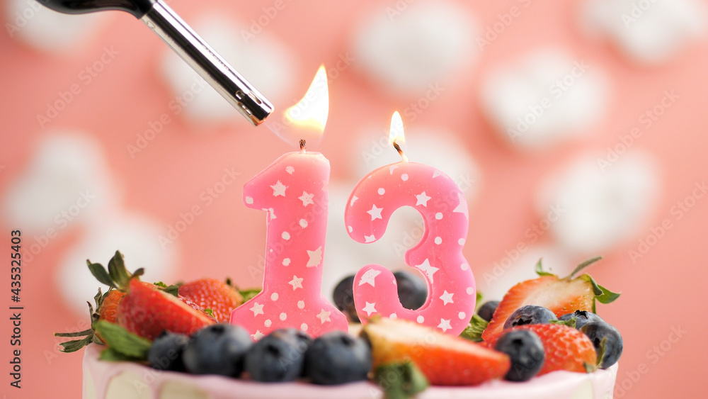 Birthday cake number 13, pink candle on beautiful cake with berries and lighter with fire against background of white clouds and pink sky. Close-up