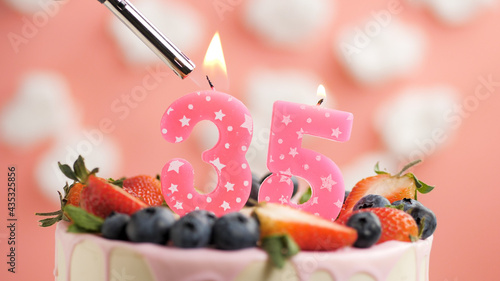Birthday cake number 35, pink candle on beautiful cake with berries and lighter with fire against background of white clouds and pink sky. Close-up