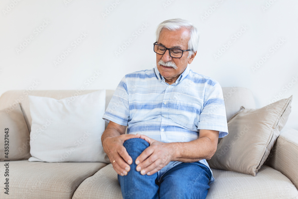 Elderly man sitting on a sofa at home and touching his painful knee. people, health care and problem concept - unhappy senior man suffering from knee ache at home Elderly man sitting on a sofa at home and touching his painful knee. people, health care and problem concept - unhappy senior man suffering from knee ache at home