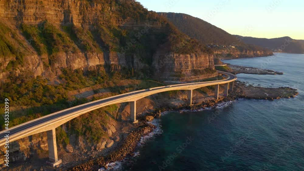 Cars Driving By The Sea Cliff Bridge In NSW Australia In Sunrise - aerial shot Cars Driving By The Sea Cliff Bridge In NSW Australia In Sunrise - aerial shot