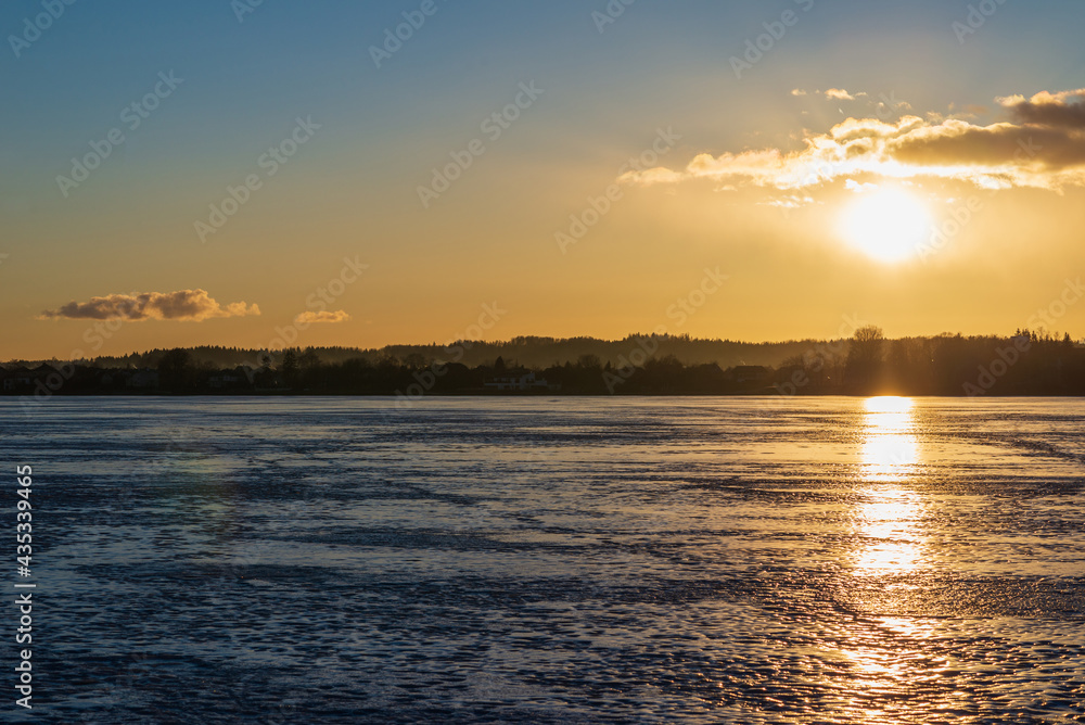 Winter landscape with frozen lake at sunrise or sunset. Lake glistening ice reflect a sun.Forest in the background.