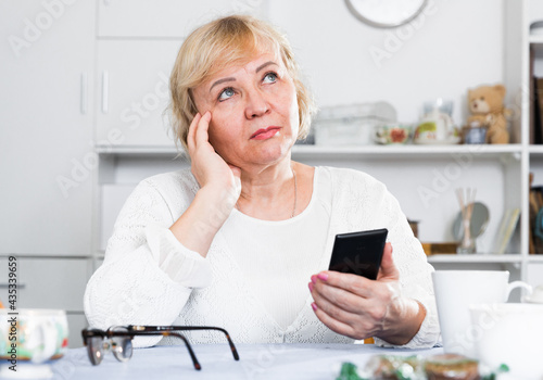 Mature woman sitting at table at home and looking at smartphone in her hands.