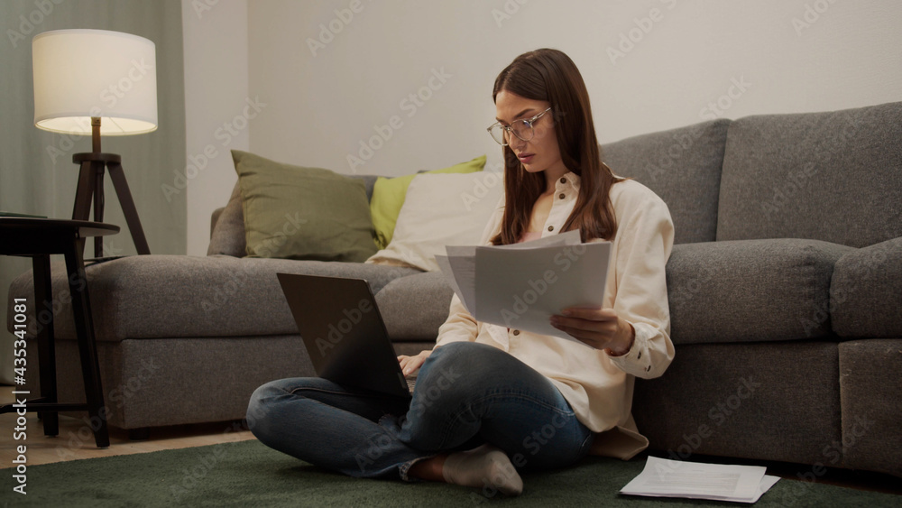 A serious woman is sorting through papers and typing on a laptop.