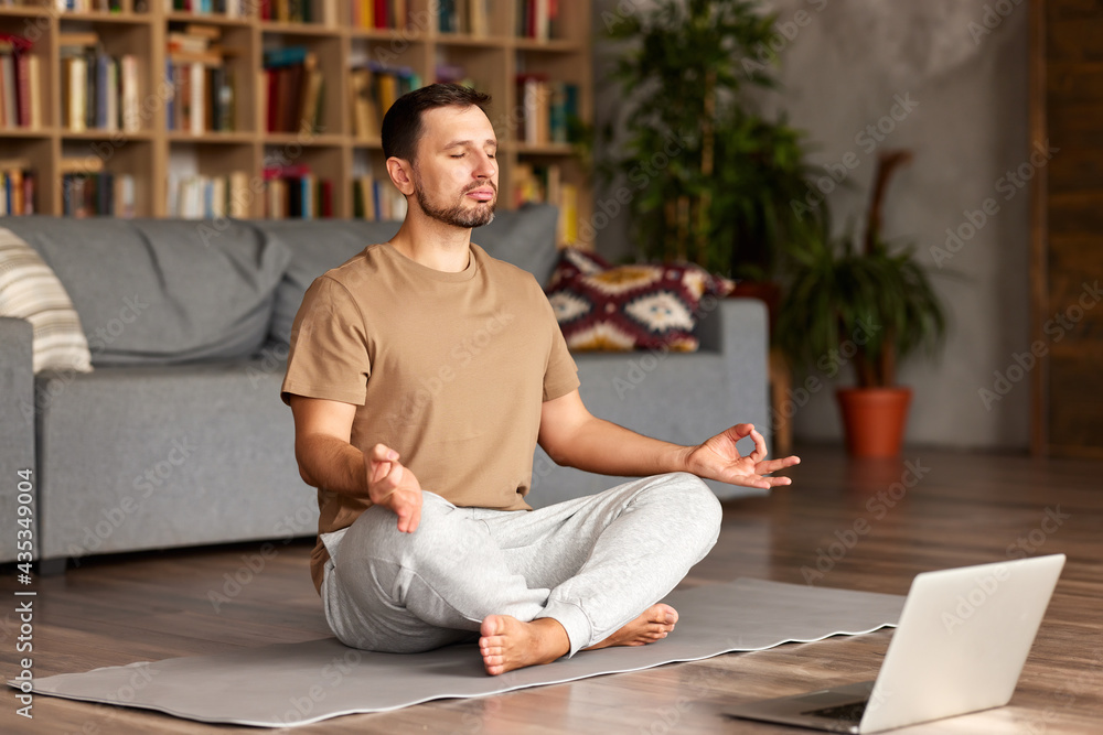 Fototapeta premium Peaceful young man meditating while sitting in lotus pose on floor at home in front of laptop