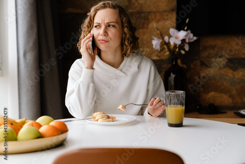 Young white woman talking on mobile phone while having breakfast at home
