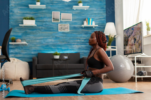 Black woman doing pilates workout using elastic strap sitting on yoga mat, pulling training arms and shoulders in home living room. Athletic fit exercising body using resistance band. photo