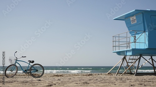Blue bicycle, cruiser bike by ocean beach, pacific coast, Oceanside California USA. Summertime vacations, sea shore. Vintage cycle on sand near lifeguard tower or watchtower hut. Sky and water waves.