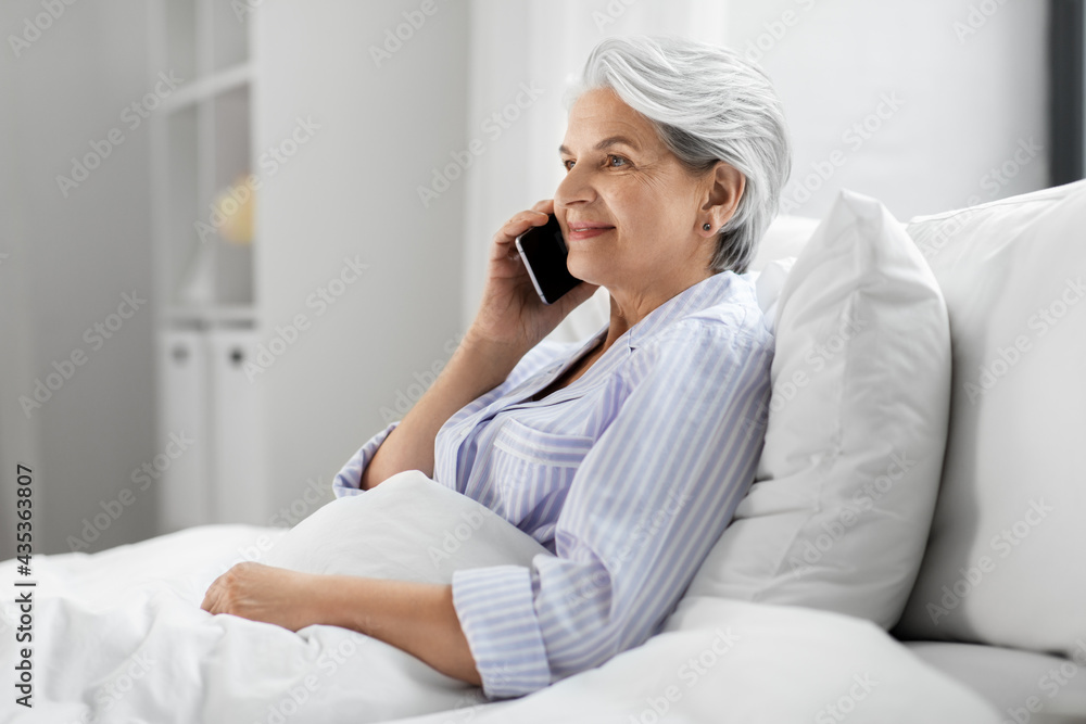 old age, technology and people concept - happy smiling senior woman in pajamas calling on smartphone sitting in bed at home bedroom old age, technology and people concept - happy smiling senior woman in pajamas calling on smartphone sitting in bed at home bedroom