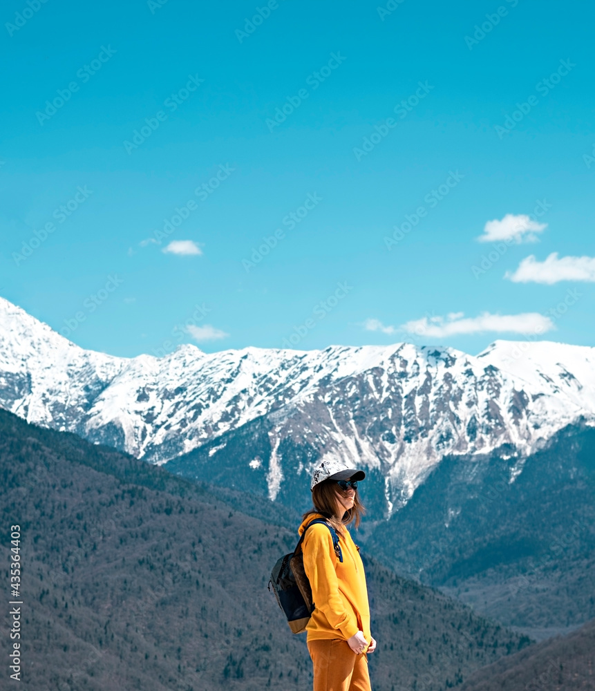 side view Young blond woman in yellow hoodie, cap, with backpack on background of snow capped Caucasus mountains. Hiking and travel, active lifestyle, copy space