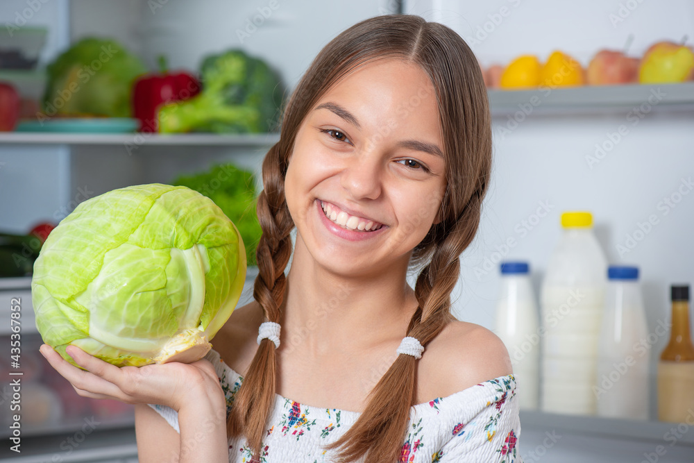 Beautiful young teen girl holding fresh green cabbage while standing near open fridge in kitchen at home Beautiful young teen girl holding fresh green cabbage while standing near open fridge in kitchen at home