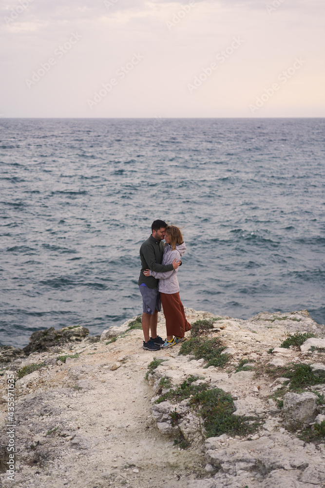 Couple is standing on a rock by the sea, hugging, enjoying the view and fresh air.