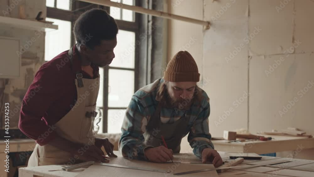 Medium shot of young African man standing at workbench and watching bearded Caucasian carpenter drawing blueprint of wooden plank using pencil and ruler Medium shot of young African man standing at workbench and watching bearded Caucasian carpenter drawing blueprint of wooden plank using pencil and ruler