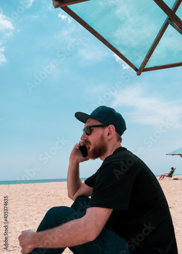 Bearded millennial man in black t-shirt snapback on blue sea landscape with smartphone in hand talking. Young adult guy calling friends on cell phone, real people communication lifestyle distance work