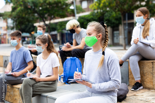 Students in protective masks record lecture while sitting on a stone street parapet
