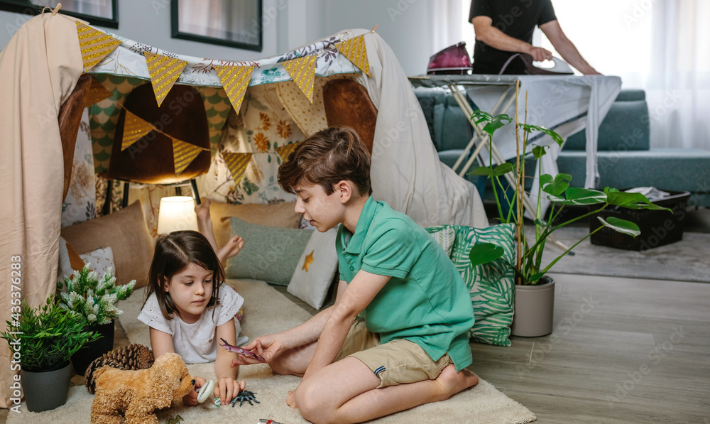 Children playing camping at home while their father irons in the living room