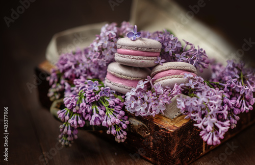 Macaroni cakes lying on a bouquet of lilacs in a wooden box on a dark wooden background.