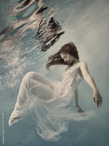 A girl with long dark hair in a white dress with glitters floats underwater as if floating in zero gravity on a light background