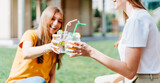  Two beautiful young women sitting on the grass and laughing while having a lemonade cocktails drink in glass jars during a friendly hang out in city park. Summer vacation and picnic at sunny day.