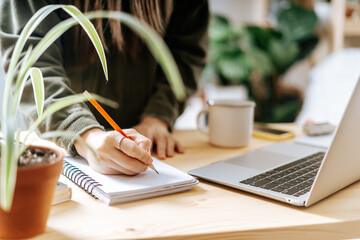 Distance learning online education and work. Business woman with plants writes in notebook. Cropped freelance girl with laptop at home office. Using computer and online shops.