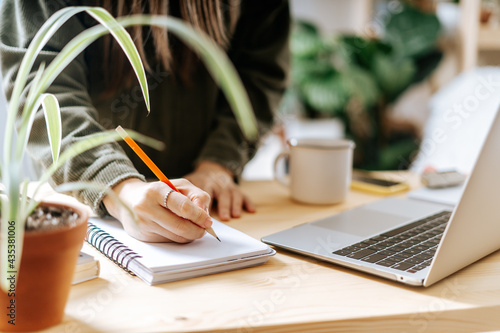 Distance learning online education and work. Business woman with plants writes in notebook. Cropped freelance girl with laptop at home office. Using computer and online shops.