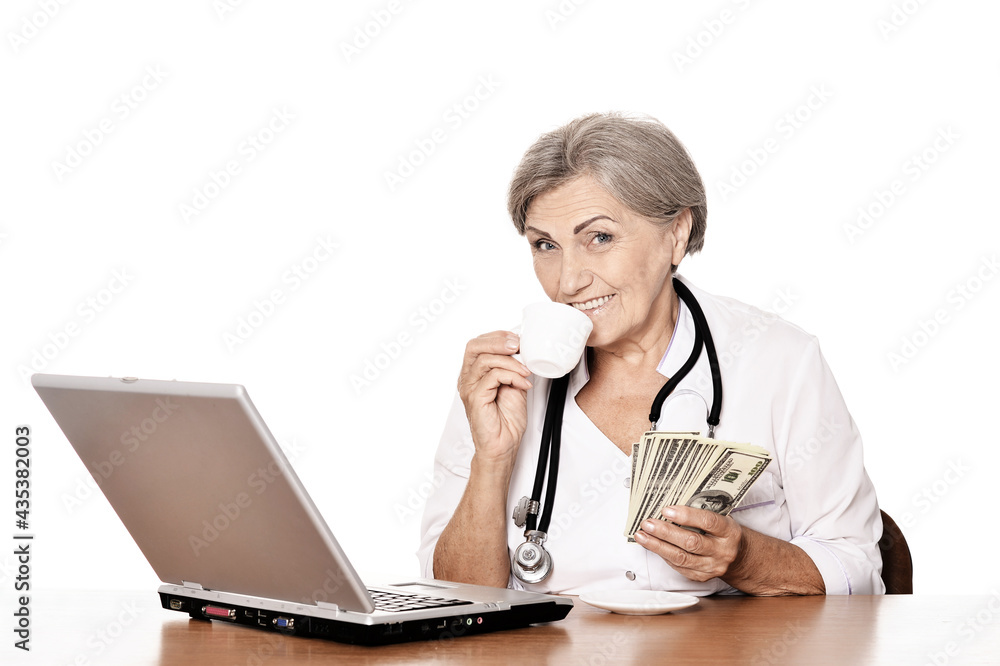 Senior doctor sitting at table with laptop and holding dollars Senior doctor sitting at table with laptop and holding dollars