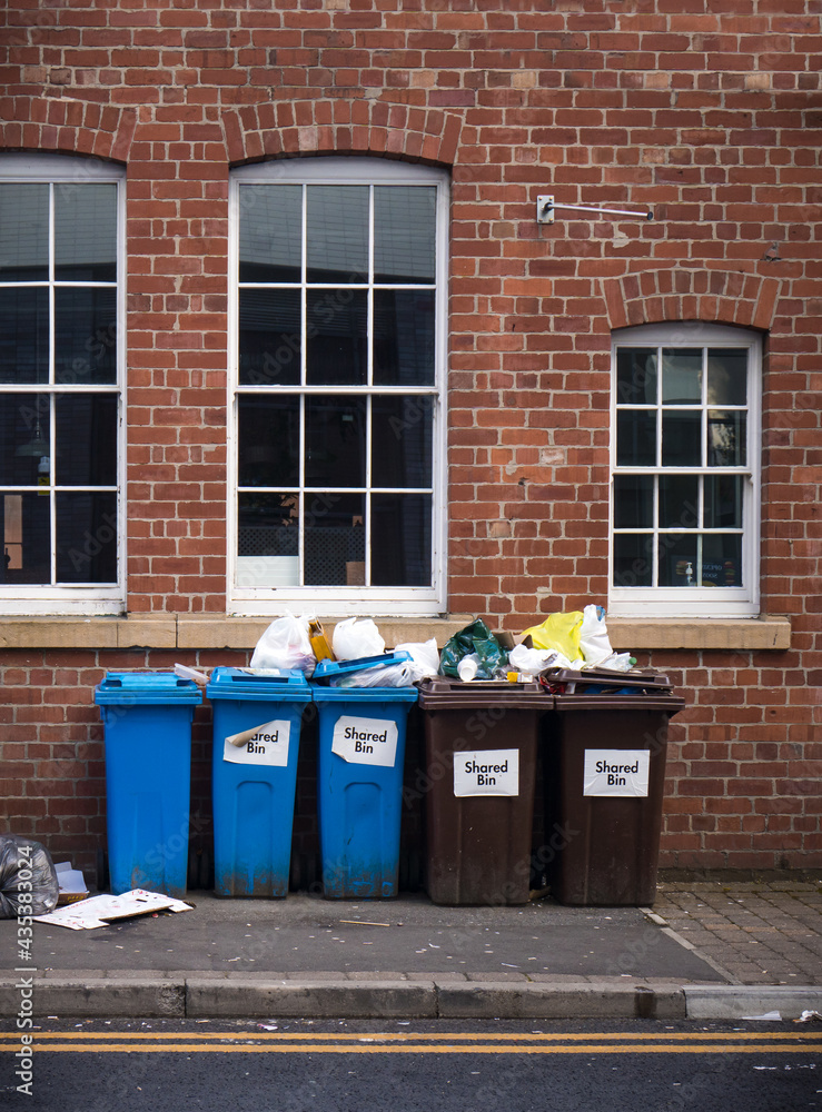 Overflowing row of rubbish bins stood on pavement in city centre. Brown