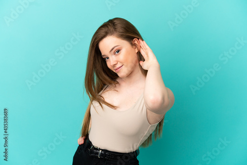 Teenager girl over isolated blue background listening to something by putting hand on the ear