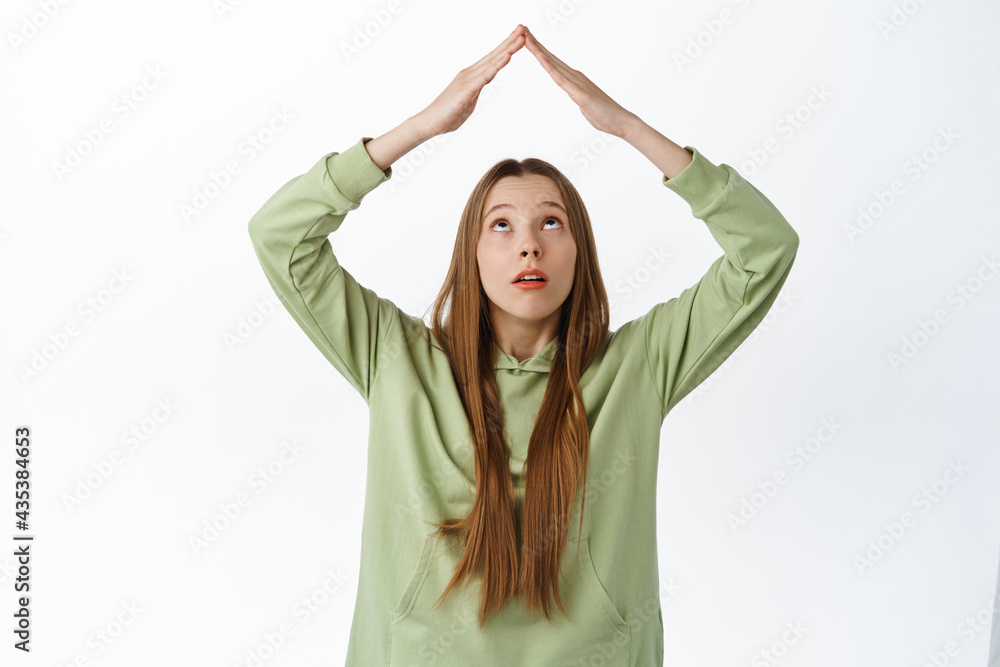 Girl looks curious at hands making rooftop gesture, show roof sign above head and staring at top, standing in hoodie against white background
