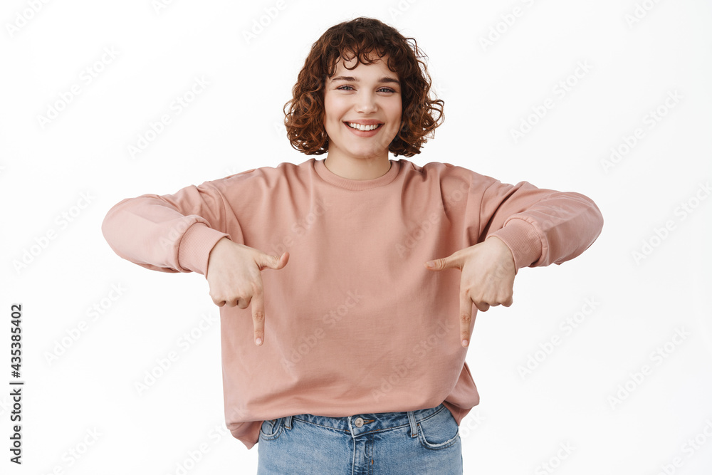 Logo below. Smiling young woman with curly hair, white teeth, showing advertisement, pointing fingers down at promo banner, standing against white background Logo below. Smiling young woman with curly hair, white teeth, showing advertisement, pointing fingers down at promo banner, standing against white background