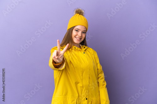 Teenager girl wearing a rainproof coat over isolated purple background smiling and showing victory sign