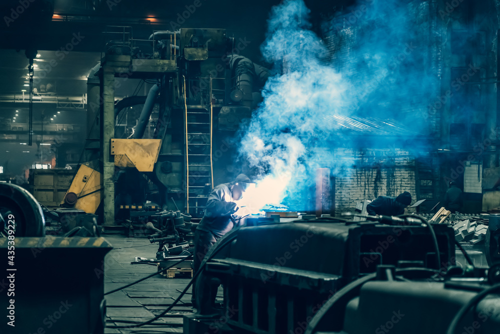 Welder in protective mask works in metallurgical factory workshop, sparks and smoke from welding flares, industrial background.