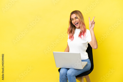 Young woman sitting on a chair with laptop over isolated yellow background smiling and showing victory sign © luismolinero