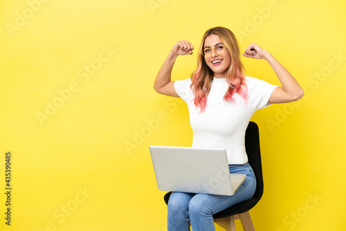 Young woman sitting on a chair with laptop over isolated yellow background doing strong gesture © luismolinero