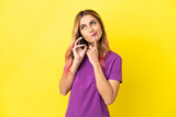 Young woman using mobile phone over isolated yellow background having doubts while looking up