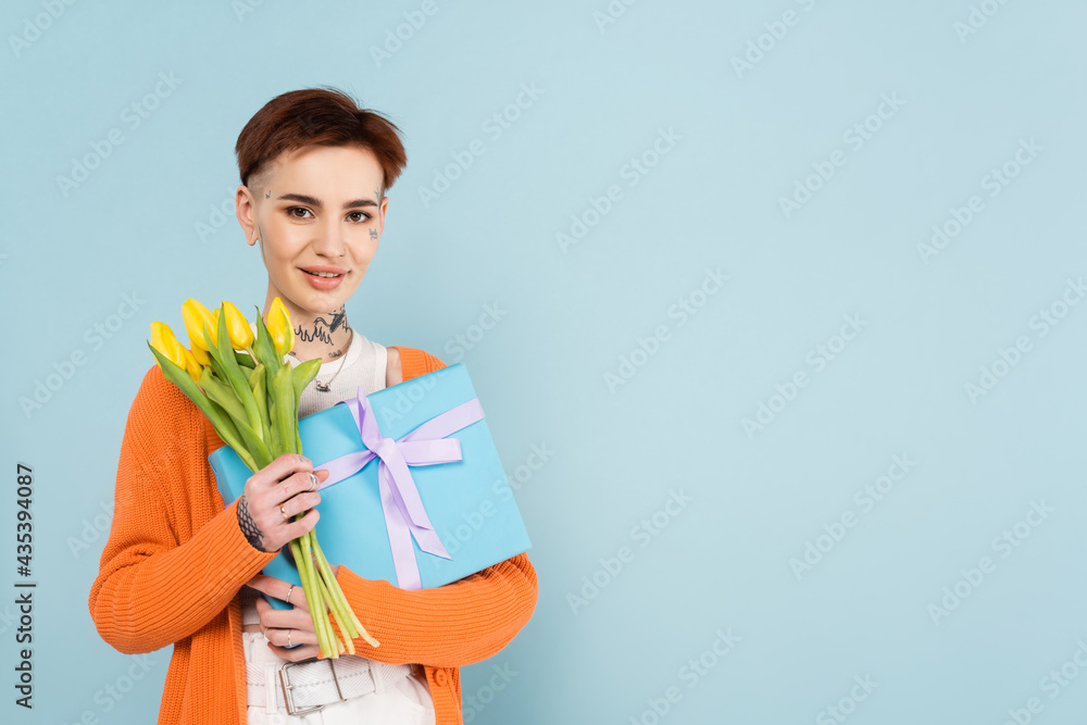 cheerful woman with tattoos holding yellow tulips and wrapped gift box isolated on blue cheerful woman with tattoos holding yellow tulips and wrapped gift box isolated on blue