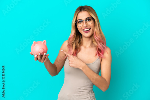 Quadro em tela Young woman over isolated blue background holding a piggybank and pointing it
