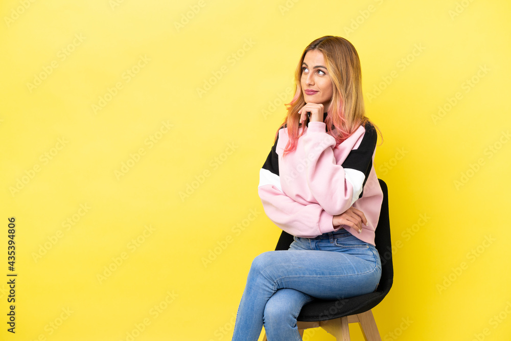Young woman sitting on a chair over isolated yellow background having doubts while looking up Young woman sitting on a chair over isolated yellow background having doubts while looking up
