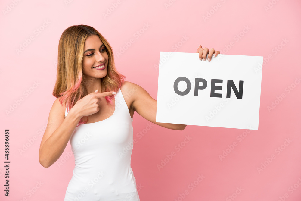 Young woman over isolated pink background holding a placard with text OPEN and pointing it Young woman over isolated pink background holding a placard with text OPEN and pointing it