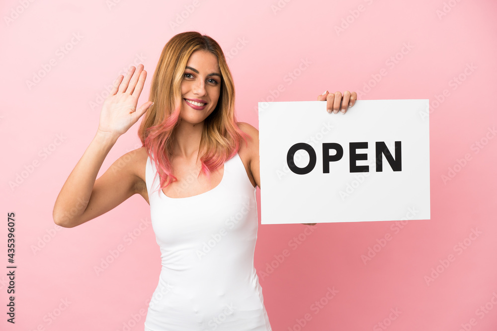 Young woman over isolated pink background holding a placard with text OPEN and saluting Young woman over isolated pink background holding a placard with text OPEN and saluting